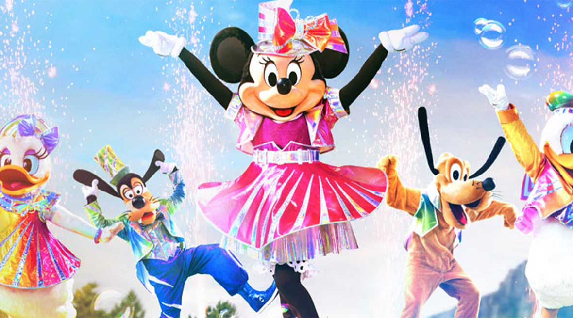 Disney characters Minnie Mouse, Donald Duck, Daisy Duck, Goofy and Pluto dance and wave in front of a crowd of people in face coverings.